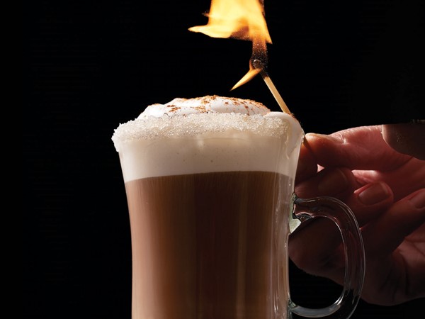 Glass of coffee topped with foam, whipped topping and cinnamon with a person holding a match above the glass