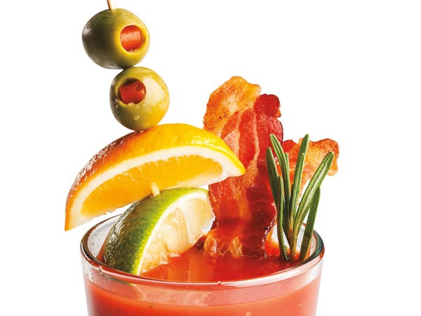 Glass filled with bloody mary, garnished with bacon slices, lime and orange wedges and stuffed olives