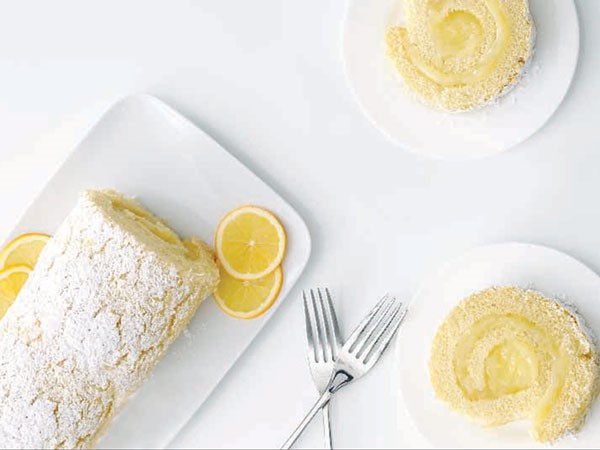 Platter of lemon cake roll with lemon slices and two forks and plates with slices of cake