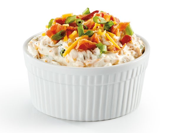Small white bowl of dip topped with shredded cheddar cheese, bacon bits and chopped green onion
