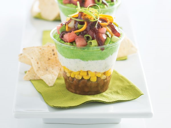 Plastic cup layered with beans, corn, sour cream, guacamole, tomatoes, green onion and cheese