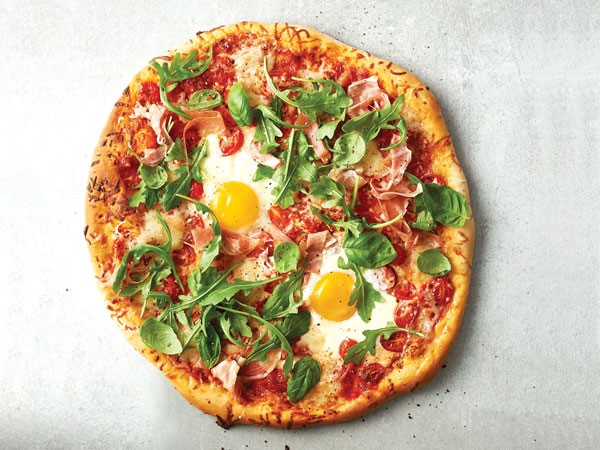 Pizza crust topped with cherry tomatoes, prosciutto, shredded pizza cheese, eggs and arugula
