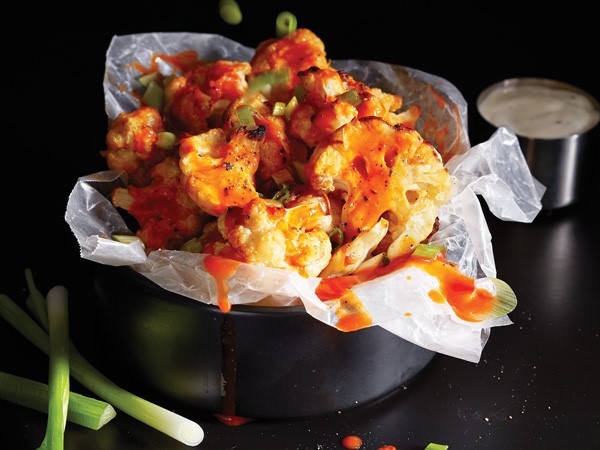 Wings in a black bowl layered with wax paper and cauliflower drenched in buffalo sauce