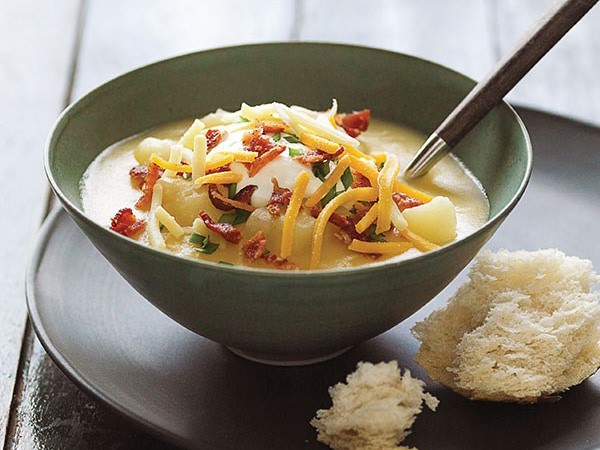 Bowl of potato soup garnished with a dollop of sour cream, shredded cheese, bacon bits and chive and served with bread