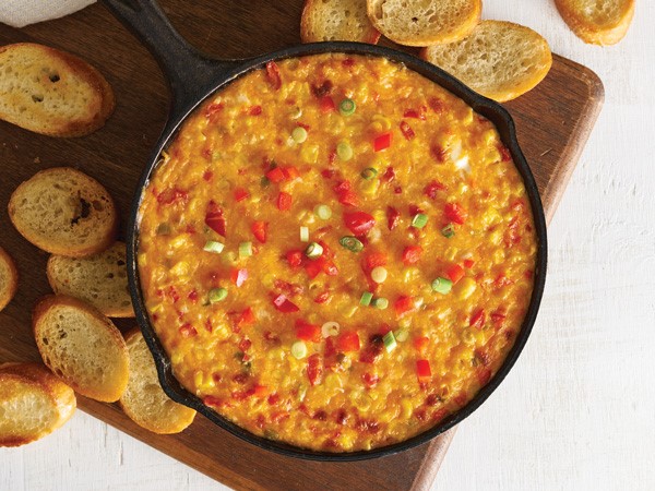 Cast-iron skillet filled with cheese dip topped with sliced green onions, diced red peppers on a wooden cutting board surrounded by sliced baguetts