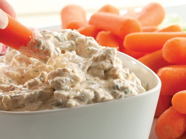 Hand dipping a baby carrot into bowl of blue cheese veggie dip, surrounded by a plate of baby carrots