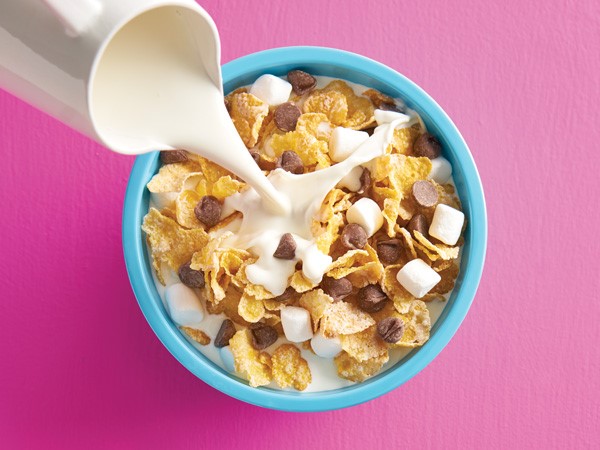 Flaked corn cereal with mini marshmallows and chocolate chips with milk being poured into blue bowl