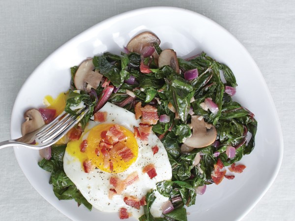 Plate of sauteed chard and eggs, garnished with red onion, mushrooms and bacon crumbles