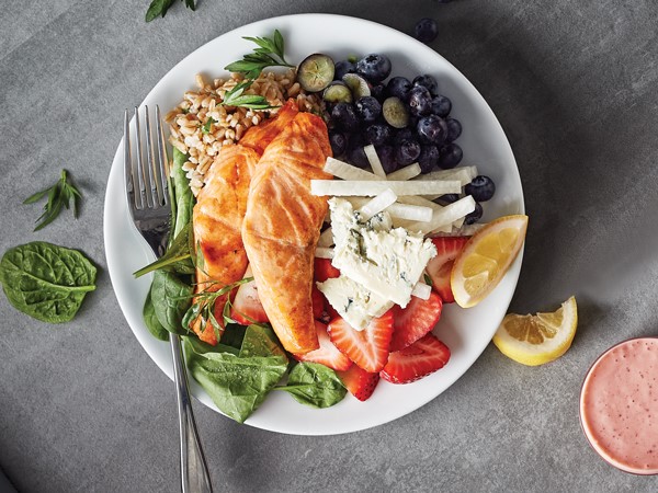 White plate filled with spinach, sliced strawberries, blueberries, farro, sliced jicama, two pieces of salmon, and sliced Gorgonzola cheese