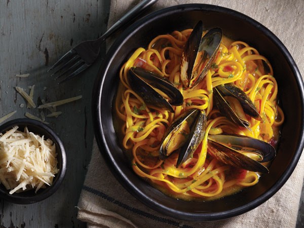 Black bowl of linguine topped with mussels and served with a small bowl of shredded parmesan