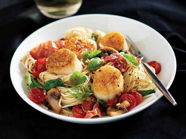 Bowl of angel hair pasta topped with seared scallops, chopped prosciutto, grape tomatoes, mushrooms, garlic and spinach and garnished with fresh basil leaves