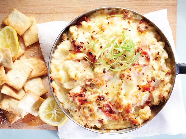 Bowl of artichoke king crab dip garnished with green onions and surrounded by crackers on a wooden board