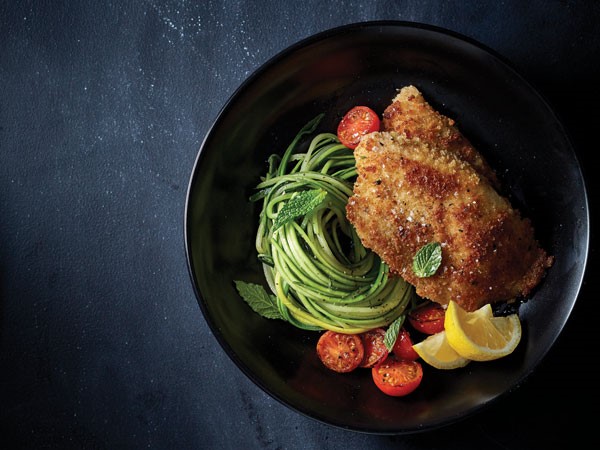 Breaded and seasoned tilapia next to fresh zucchini pasta, blistered and halved cherry tomatoes with fresh slices of lemon and mint leaves for garnish