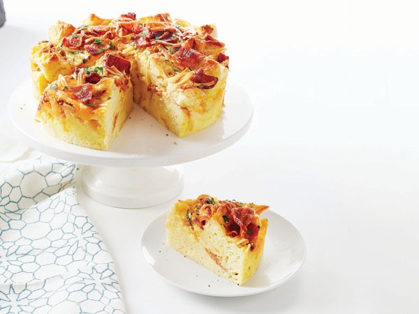 Round strata filled with bread, eggs, bacon, and melted cheese on white cake stand