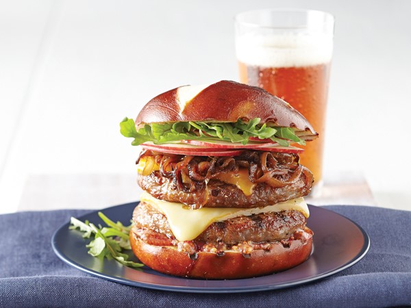Two burger patties separated by a slice of cheese topped with caramelized onions, thinly sliced red apples and arugula on a pretzel bun
