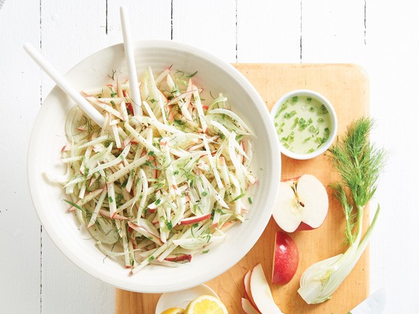 Small bowl of crunchy apple and fennel slaw next to cutting board with apple slices, fennel and dressing