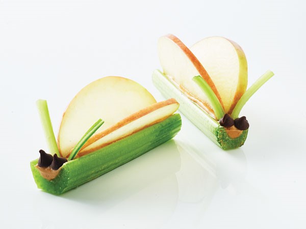 Celery sticks filled with peanut butter and topped with two chocolate chips, two apple slices and two thin strips of celery to form a celery critter