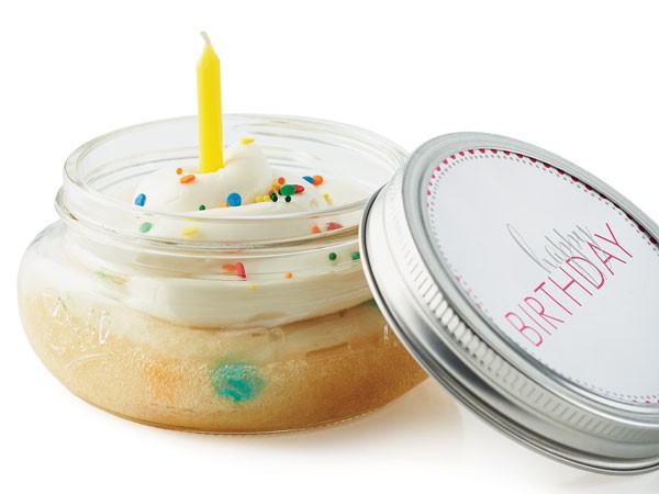 Half-pint-size mason jar filled with cake and topped with white frosting with colored sprinkles and a yellow birthday candle