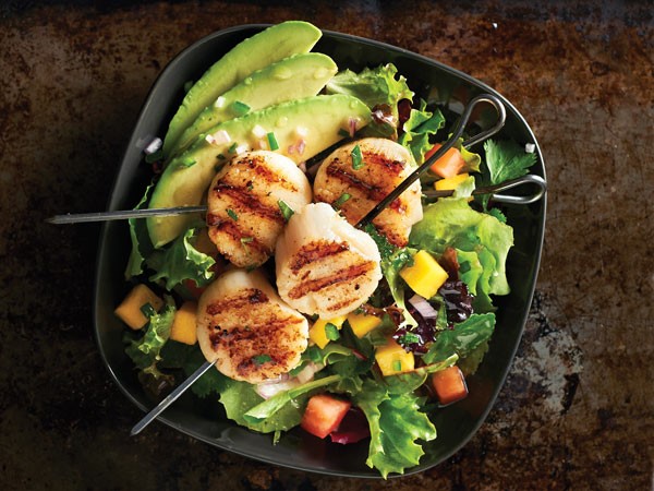 Grilled scallops on skewer in dark bowl with avocado and salad greens