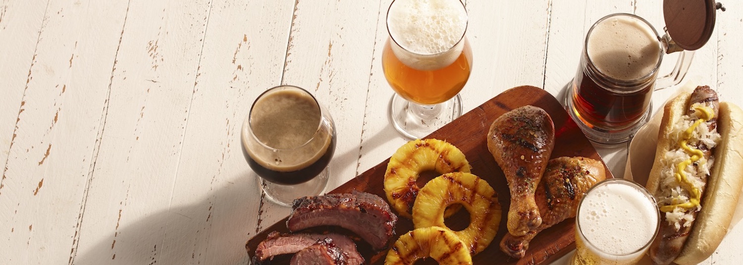 White wooden background with beer, brats, grilled pineapple, chicken, and ribs