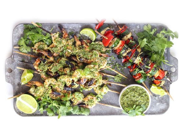 Platter of grilled shrimp covered in chimichurri sauce on skewers next to skewered vegetables