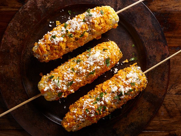 Skewered corn on the cob topped with sour cream, crushed nacho chips, cotija cheese and fresh cilantro on a brown plate