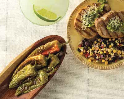 Grilled whole hatch chile peppers next to a plate of pork chops and bean salsa