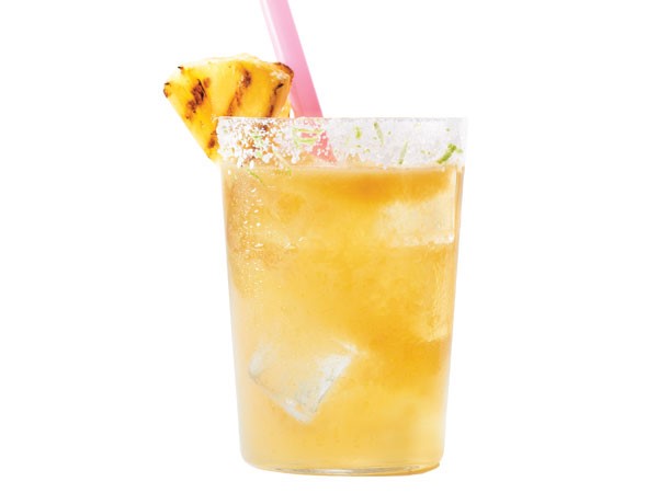 Sugar-rimmed glass of pineapple margarita, garnished with a pink straw and grilled pineapple wedge