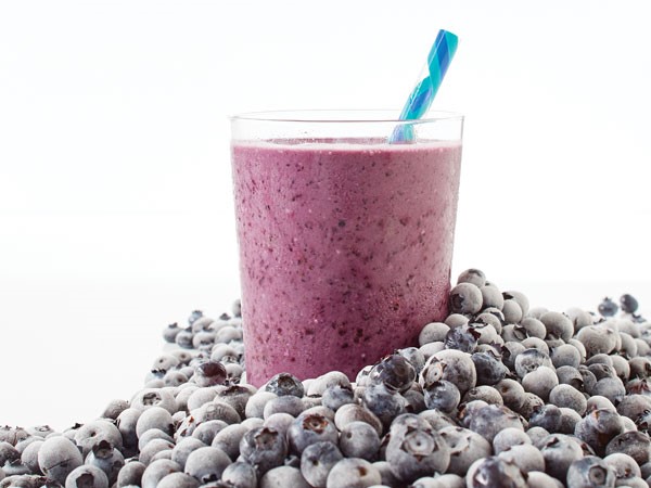 Glass of blueberry oatmeal smoothie with a blue straw, surrounded by fresh blueberries