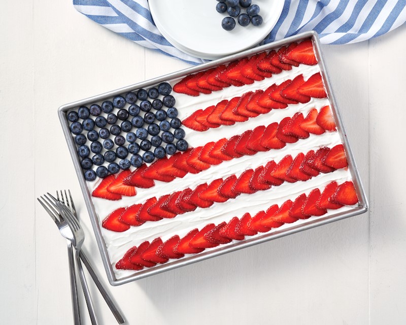 Flag pudding cake in a 13x9 baking pan topped with white frosting and decorated like an American flag using berries for stars and slices of strawberries for stripes.