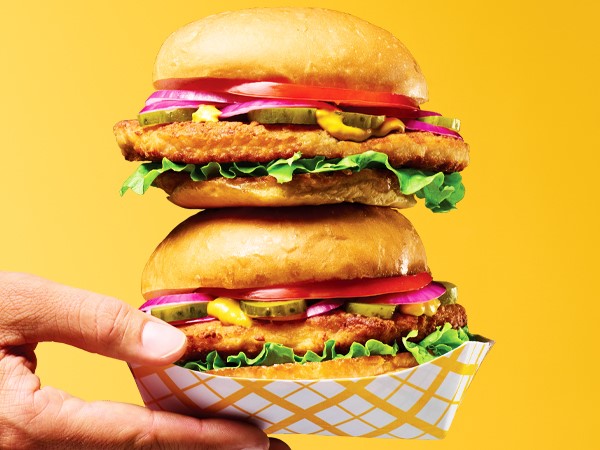 Two stacked breaded pork loin sandwiches in white and yellow takeout boats held in a hand on a yellow background. 