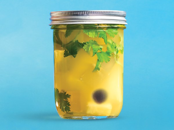 Mason jar filled with pineapples and cilantro in a yellow-colored liquid on a blue background. 