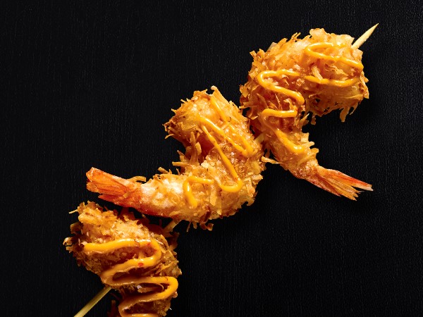 3 fried shrimp on a stick drizzled with sauce on a black background. 