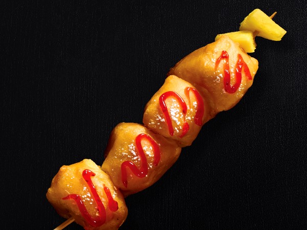 Fried pineapple chunks on a stick drizzle with cherry sauce and sprinkled with powdered sugar on a black background.