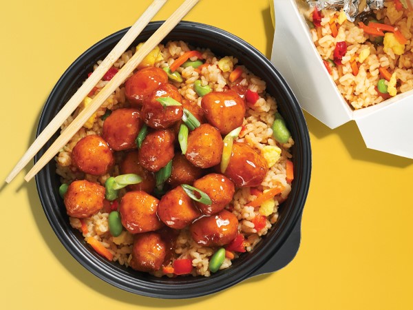 A black plastic bowl filled with orange chicken and fried rice, topped with chopsticks, and next to a white take out box on a yellow background. 