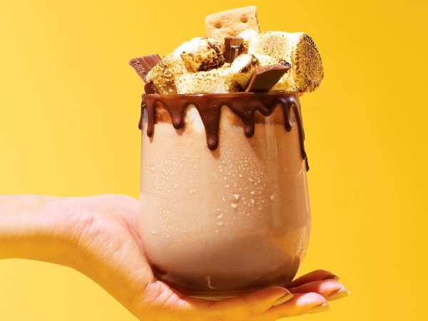 A glass of s'more flavored wine topped with marshmallows, chocolate, and graham crackers. Melted chocolate drips from the rim of the glass that sits in the palm of a hand on a yellow background. 
