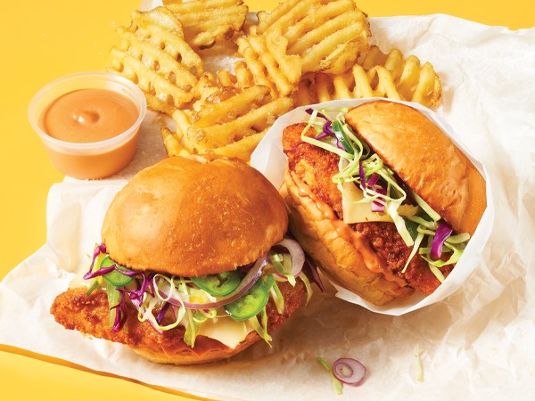 Two fried chicken sandwiches on a napkin with waffled French fries and dipping sauce on a yellow background.