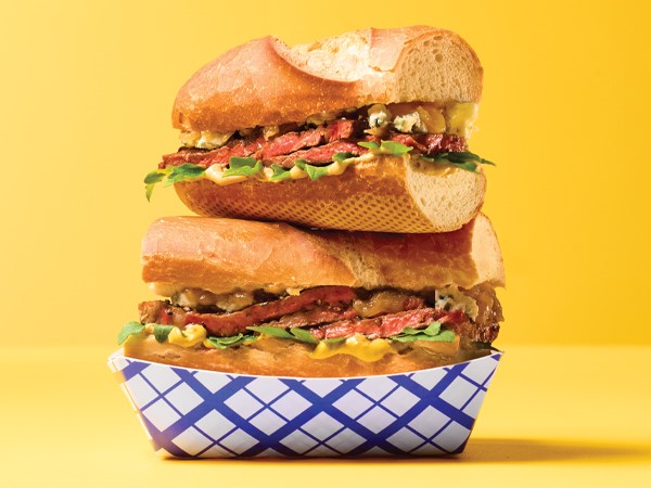 Two ribeye steak sandwiches stacked in a white and blue takeout boat on a yellow background.