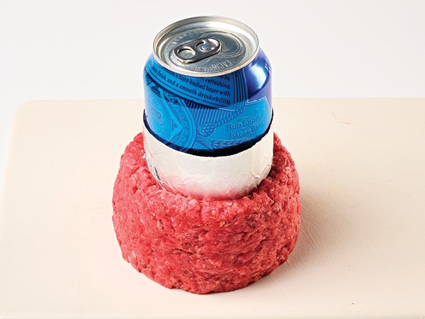 Ground beef formed around a foil-wrapped blue beer can on a white background.