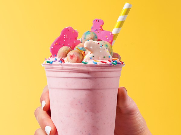 Pink-colored frosted animal cookie shake topped with frosted animal cookies, edible cookie dough bites, rainbow sprinkles, and a yellow-and-white =-striped straw. Held in a female hand with white nail polish on a yellow background.