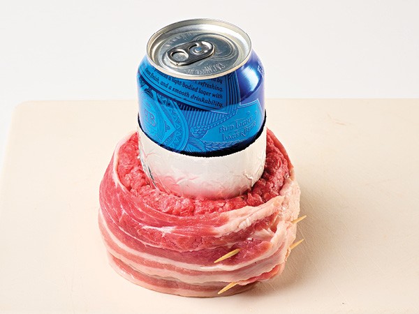Bacon-wrapped ground beef formed around a foil-wrapped blue beer can on a white background. 