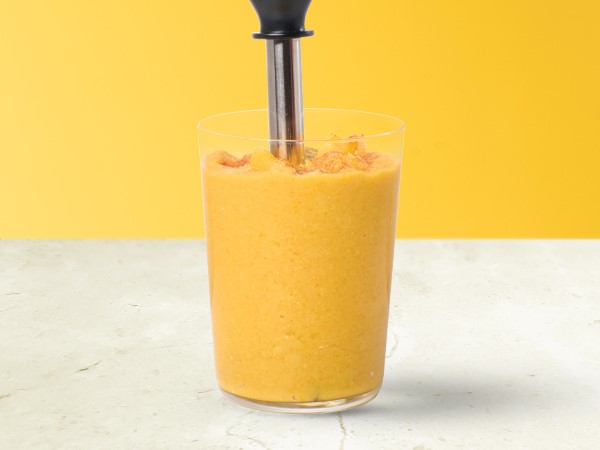 Ginger-peach hummus in a glass with an immersion blender on a white and yellow background. 
