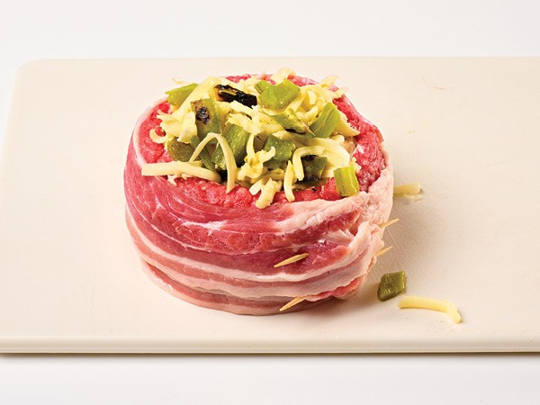 Bacon-wrapped ground beef filled with cheeses and peppers on a cream cutting board.