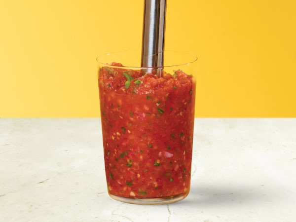Tomato-basil salsa in a glass with an immersion blender inserted in the salsa on a white and yellow background.