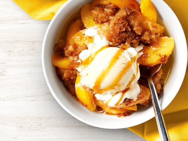 A white bowl with peach crumble topped with vanilla ice cream and caramel sauce with a spoon. Bowl sits on a light cream background and a yellow linen. 