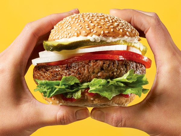 A plant-based burger topped with lettuce, ketchup, tomato slices, onion slices, pickle slices, and mayonnaise on a sesame seed bun, held with two hands on a yellow background.