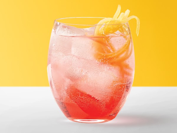 Red spritzer in an ice-filled glass garnished with lemon peels on a white and yellow background.
