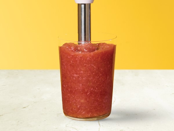 Strawberry applesauce in a glass with an immersion blender in the applesauce on a white and yellow background. 