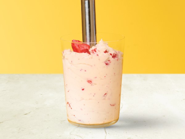 Strawberry whipped cream in a glass with an immersion blender in the whipped cream on a white and  yellow background. 