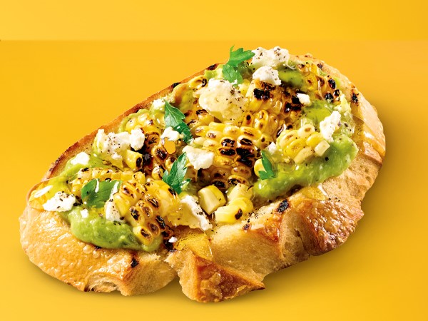 Grilled sweet corn, avocado, and feta topped on a grilled piece of sourdough bread on a yellow background. 